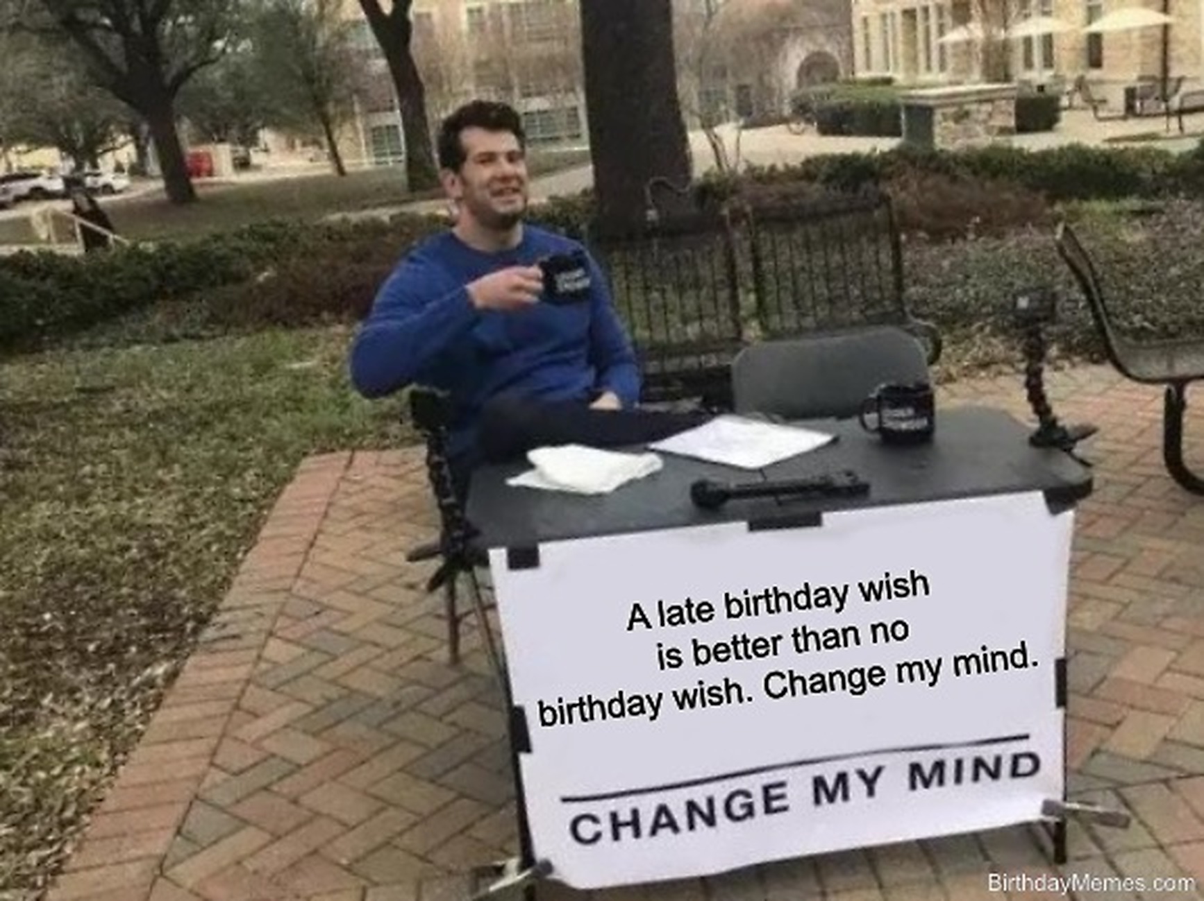 Man at a table with a sign reading 'A late birthday wish is better than no birthday wish. Change my mind.'