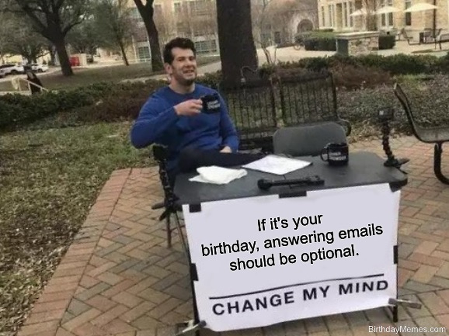 A man sits at a table outdoors with a sign that says, 'If it's your birthday, answering emails should be optional. Change my mind.'